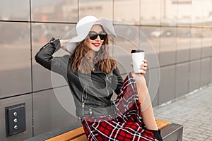 Pretty positive woman with sweet smile in vintage sunglasses in hat in casual red-black clothes sit on bench with coffee and