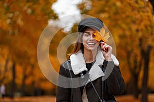 Pretty positive young woman in a fashionable black hat in a stylish brown  jacket holds a yellow-orange maple leaf near face and