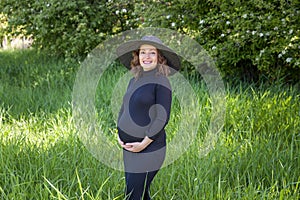 Pretty pregnant woman in a black dress and hat green field park