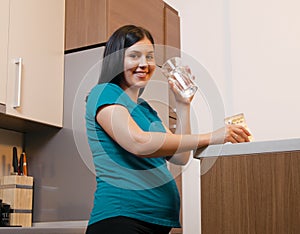 Pretty pregnant woman drinking glass of water in the kitchen, holding vitamins