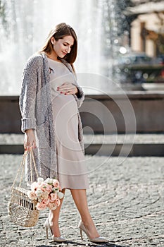 Happy pregnant woman with flowers walking near fountain