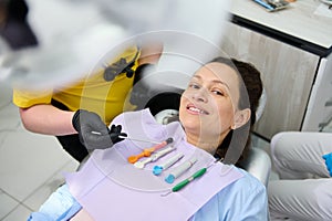 Pretty woman in dentist's chair with dental instruments and anesthetics on napkin on her chest, smiles looking at photo