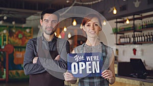 Pretty red-haired businesswoman cafe owner is holding `yes we are open` sign with her employee in apron standing near