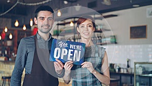 Pretty red-haired businesswoman cafe owner is holding Yes We Are Open sign with her employee in apron standing near her