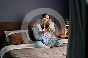 Pretty redhead young woman posing for mobile selfie in bed while sitting on bed at cozy bedroom