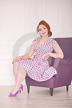 Pretty redheaded pin up woman wearing pink polka dot dress and posing with purple armchair on white background
