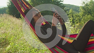Pretty relaxed african american woman lying in hammock,networking online on phone in mountains