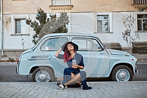 Pretty relaxing woman in hat sitting near vintage blue car. Lady with cup of drink. Leisure time, coffee break concept