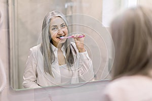 Pretty retired woman with long gray hair, wearing white shirt, brushing her teeth in the bathroom at home, standing in