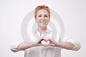 Pretty romantic mature redhead woman making a heart gesture with her fingers in front of her chest showing her love