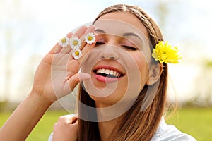 Pretty romantic young woman with flower on ear and daisies between fingers, staying outdoors with closed eyes, close up portrait
