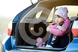 Pretty sad child girl sitting alone in a car trunk with a pink toy teddy bear