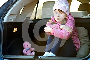 Pretty sad child girl sitting alone in a car trunk with a pink toy teddy bear