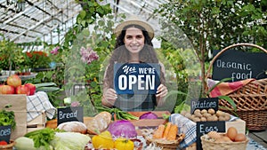 Pretty saleswoman holding open sign in farm market smiling looking at camera