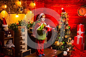 Pretty Santa boy holds New Year gift box. Happy new year. Smiling little boy in Santa helper hat over christmas tree at