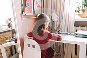 Pretty school girl with long hair in red dress draws with pencil at table in childrens room at home