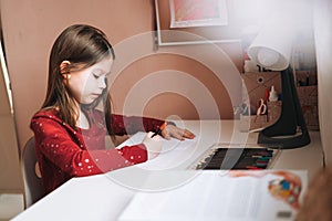 Pretty school girl with long hair in red dress draws with pencil at table in childrens room at home