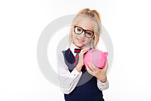 Pretty schoolgirl with blonde hair in school suit smiles and holds pink pig money box with money isolated on white