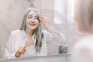 Pretty senior woman with long gray hair, wearing white shirt, looking at her face in the bathroom mirror, and applying