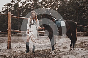 Pretty and sensual young woman wearing the dress is holding the reins and posing with the horse on the ranch