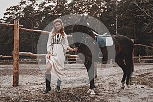 Pretty and sensual young woman wearing the dress is holding the reins and posing with the horse on the ranch photo
