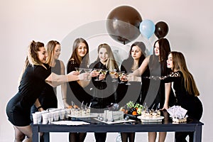 Pretty seven girls in black dresses touching the martini glasses with each other, celebrating birthday, standing near