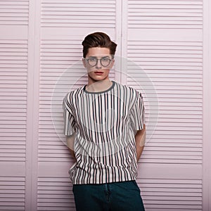 Pretty sexy young man hipster in fashionable glasses with a hairstyle in a trendy striped t-shirt stand near a vintage wooden pink