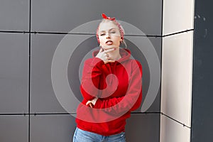 Pretty sexy young woman with blond hair in a fashionable red sweatshirt with a hood in a stylish bandana in vintage jeans poses