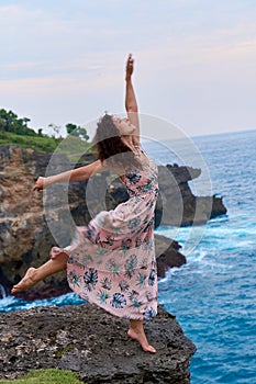 A pretty slender woman in a long dress poses on a rock on a cliff above the ocean