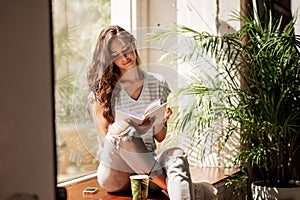 A pretty slim young girl with long hair,wearing casual outfit,sit on the windowsill and reads a book in a cozy cafe.