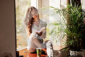 A pretty slim young girl with long hair,wearing casual outfit,sit on the windowsill and reads a book in a cozy cafe.