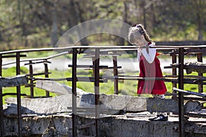 Pretty small blond long haired girl in nice red dress stands alone on old cement bridge leaning on wooden railings looking intent