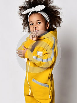 Pretty small curly mulatto african girl in warm yellow jumpsuit and hair bow standing and showing silence sign