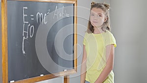 Pretty smart Caucasian schoolgirl posing at chalkboard with physical formulas written. Portrait of cute confident genius