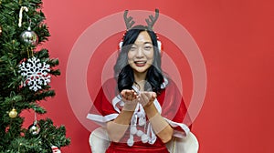 Pretty smiling asian woman in santy costume and santa hat is making a wish next to christmas tree on red background for season