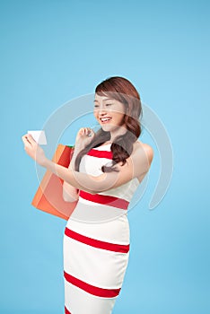 Pretty smiling Asian woman with shopping bags showing credit card in hand studio shot isolated on blue background