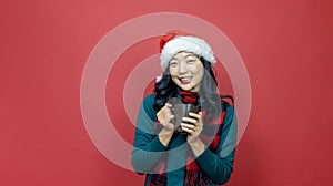 Pretty smiling asian woman in warm christmas sweater and santa hat is having hot chocolate on red background for season