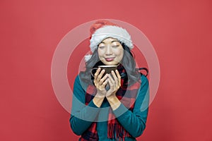 Pretty smiling asian woman in warm christmas sweater and santa hat is having hot chocolate on red background for season