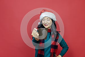 Pretty smiling asian woman in warm christmas sweater and santa hat is having hot chocolate on red background for season