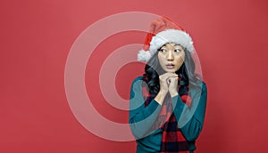 Pretty smiling asian woman in warm christmas sweater and santa hat is having skeptical idea on red background for season