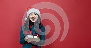 Pretty smiling asian woman in warm christmas sweater and santa hat holding gift box as present with red background for season