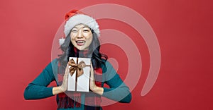 Pretty smiling asian woman in warm christmas sweater and santa hat holding gift box as present with red background for season