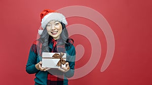 Pretty smiling asian woman in warm christmas sweater and santa hat holding gift box as present with red background for season