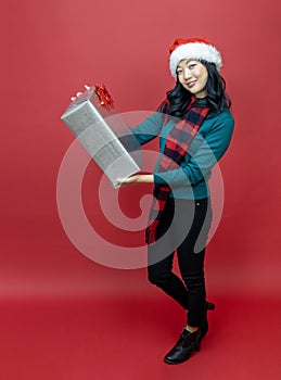 Pretty smiling asian woman in warm christmas sweater and santa hat holding gift box as present with red background for season