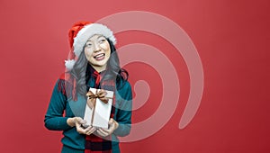 Pretty smiling asian woman in warm christmas sweater and santa hat holding gift box as present with red background for season