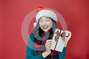 Pretty smiling asian woman in warm christmas sweater and santa hat holding gift box as present with red background for season