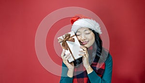 Pretty smiling asian woman in warm christmas sweater and santa hat holding gift box as present with red background for season