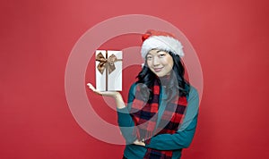 Pretty smiling asian woman in warm christmas sweater and santa hat holding gift box as present with red background for season