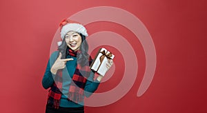 Pretty smiling asian woman in warm christmas sweater and santa hat pointing at gift box as present with red background for season