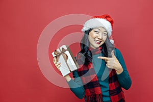 Pretty smiling asian woman in warm christmas sweater and santa hat pointing at gift box as present with red background for season